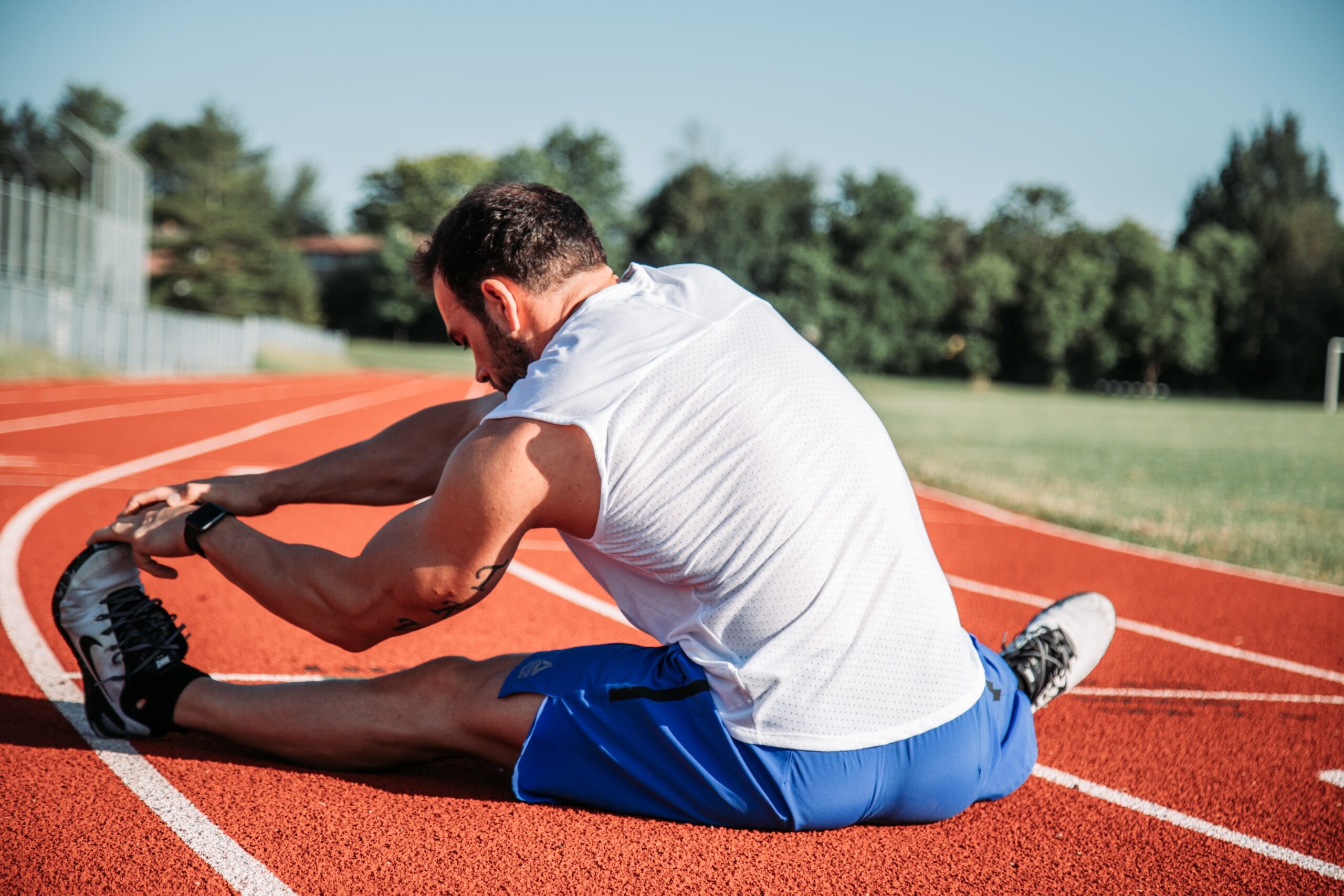 A man stretches his legs on a track and field oval, engaging in athletic conditioning under the guidance of a physiotherapist.